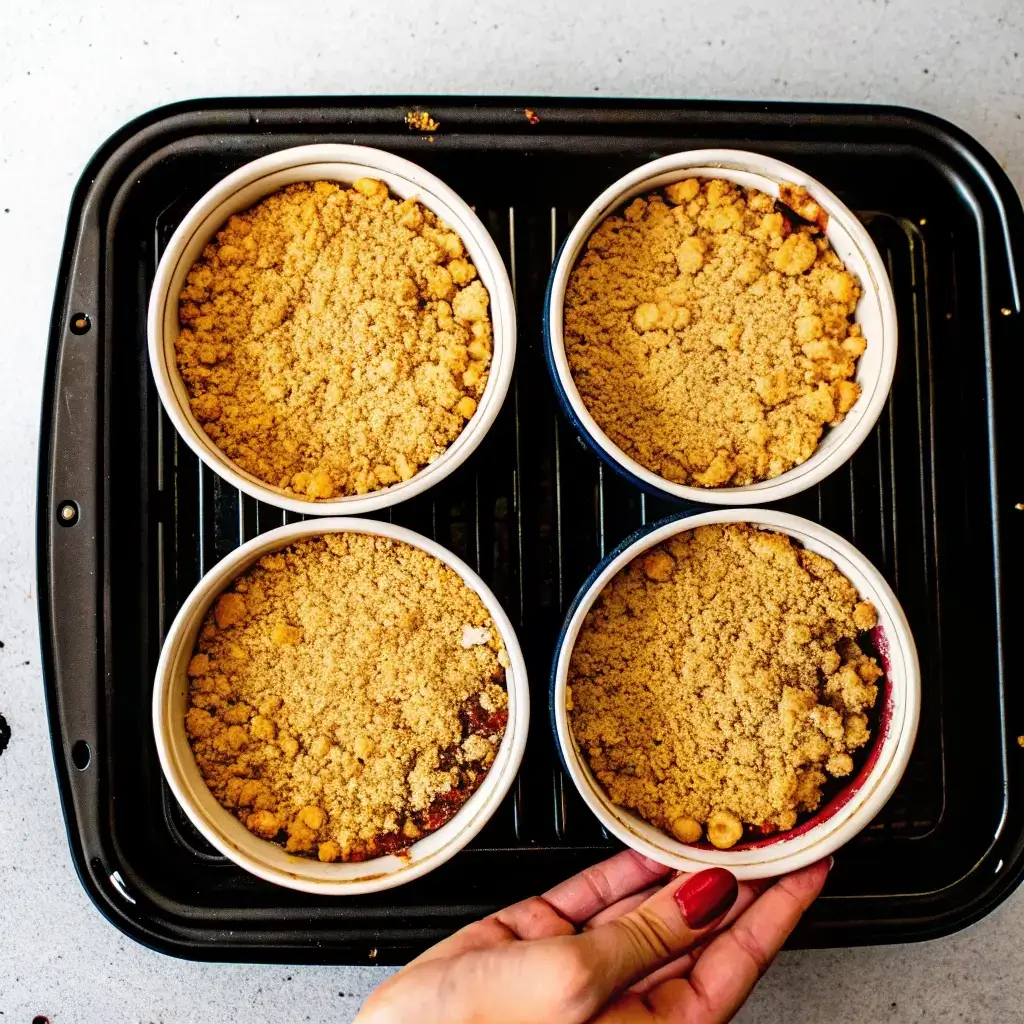 Vier Portionen Apfel Crumble in kleinen Keramikschalen, mit knusprigem Streuselbelag, auf einem Airfryer-Backblech platziert – bereit zum Backen im Airfryer. Eine Hand hält eine Schale, um die Zubereitung zu zeigen.