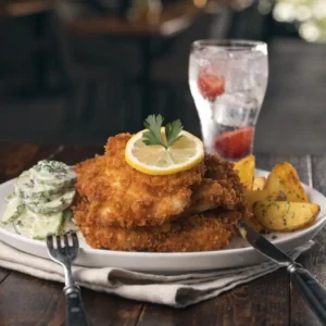 Kohlrabi Schnitzel servieren auf weißem Teller mit Zitronenscheibe, Gurkensalat und Kartoffelspalten, rustikale Tischdekoration