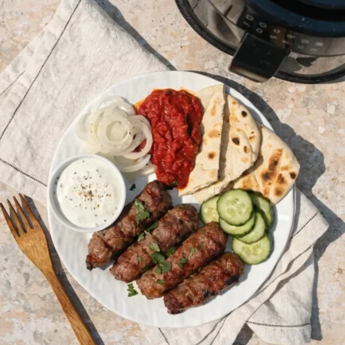 Cevapcici im Airfryer Servieren auf weißem Teller mit Ajvar, Dip, Zwiebeln, Gurken und Fladenbrot neben Airfryer-Korb